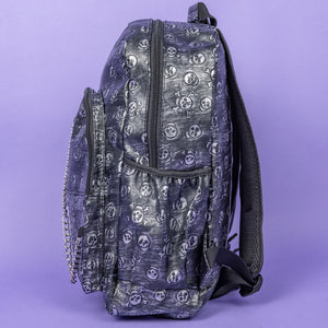The Rustic Skulls Backpack sat on a purple background. The bag is facing left to highlight the two front zip pockets, two elasticated side pockets, main zip compartment, the top handle and the detachable decorative silver chain. All over the backpack is an embossed 3d texture skulls and skull and crossbones on a faux leather material in a brushed black and silver grunge style.