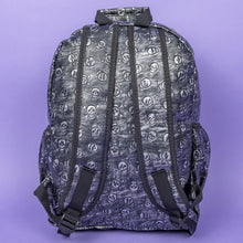 Load image into Gallery viewer, The Rustic Skulls Backpack sat on a purple background. The bag is facing away to highlight the two padded shoulder straps, two elasticated side pockets, main zip compartment and the top handle. All over the backpack is an embossed 3d texture skulls and skull and crossbones on a faux leather material in a brushed black and silver grunge style.
