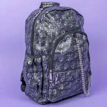 Load image into Gallery viewer, The Rustic Skulls Backpack sat on a purple background. The bag is facing forward angled slightly right to highlight the two front zip pockets, two elasticated side pockets, main zip compartment, the top handle and the detachable decorative silver chain. All over the backpack is an embossed 3d texture skulls and skull and crossbones on a faux leather material in a brushed black and silver grunge style.
