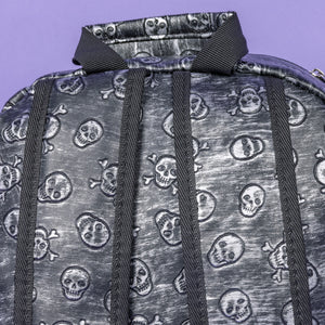 The Rustic Skulls Backpack sat on a purple background. A close up of the bag top handle and back padded shoulder straps. All over the backpack is an embossed 3d texture skulls and skull and crossbones on a faux leather material in a brushed black and silver grunge style.
