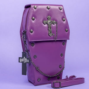 The GothX Purple Coffin Backpack. sat facing forward angled slightly right on a lilac background. The coffin bag in a sleek faux leather is a coffin shape with a magnetic close clip flap with a cross chain emblem in the centre, surrounding the coffin shape is mini metal cross studs. Draping across the front of a silver detachable decorative chain with the GothX black cross tag hanging to the left side.