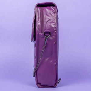 The GothX Purple Coffin Backpack. sat facing left on a lilac background. The coffin bag in a sleek faux leather is a coffin shape with a magnetic close clip flap with a cross chain emblem in the centre, surrounding the coffin shape is mini metal cross studs. Draping across the front of a silver detachable decorative chain with the GothX black cross tag hanging to the left side.