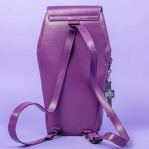 The GothX Purple Coffin Backpack is sat facing away on a lilac background. The coffin bag in a sleek faux leather is a coffin shape with a detachable adjustable strap threaded through the back. On either side are D rings for the detachable and adjustable strap, there is also a D ring and two D rings at the top to rethread the strap through to make it a backpack with the GothX black cross tag hanging to the right side.