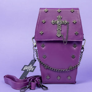 The GothX Purple Mini Coffin Bag sat facing forward on a lilac background. The coffin bag in a sleek faux leather is a coffin shape with a magnetic close clip flap with a cross chain emblem in the centre, surrounding the coffin shape is mini metal cross studs. Draping across the front of a silver detachable decorative chain with the GothX black cross tag hanging to the left side.