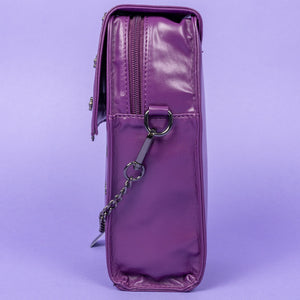 The GothX Purple Mini Coffin Bag sat facing left on a lilac background. The coffin bag in a sleek faux leather is a coffin shape with a magnetic close clip flap with a cross chain emblem in the centre, surrounding the coffin shape is mini metal cross studs. Draping across the front of a silver detachable decorative chain with the GothX black cross tag hanging to the left side.