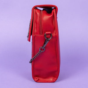 The GothX Red Mini Coffin Bag facing left to highlight the clip close magnetic flap and main zip compartment on a purple background. The coffin bag is made with vegan friendly sleek red leather and features a detachable decorative metal chain with a large metal cross and chain emblem with surrounding cross studs.