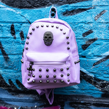 Load image into Gallery viewer, GothX Pastel Lilac Skull Head Small Studs Vegan Mini Backpack hanging on a blue graffiti wall. The vegan pastel leather bag is facing forward to highlight the metal stud detailing, the metal skull centrepiece, front zip pocket, two side pockets and top handle. The bag is inspired by pastel goth and soft grunge fashion.
