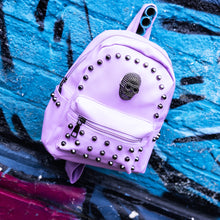Load image into Gallery viewer,  GothX Pastel Lilac Skull Head Small Studs Vegan Mini Backpack hanging on a blue graffiti wall. The vegan pastel leather bag is facing forward to highlight the metal stud detailing, the metal skull centrepiece, front zip pocket, two side pockets and top handle. The bag is inspired by pastel goth and soft grunge fashion.
