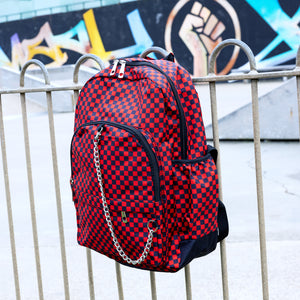 The Red Checkerboard Backpack hanging off a metal railing in a skatepark. The vegan friendly bag is facing forward to highlight the red and black check print, two front zip pockets, two elasticated side pockets, main top double zip pocket and silver draping decorative chain.