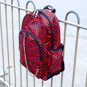 The Red Checkerboard Backpack hanging off a metal railing in a skatepark. The vegan friendly bag is facing forward to highlight the red and black check print, two front zip pockets, two elasticated side pockets, main top double zip pocket and silver draping decorative chain.