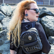 Load image into Gallery viewer, A gothic alternative blonde haired model with stretched ears and piercings wearing a black dress with black sunglasses is stood near a beach wearing the GothX black vegan skull head mini stud backpack on one shoulder with a goth outfit. The vegan leather goth bag is facing the camera showing off the handle, the skull, mini studs and front zip pocket.
