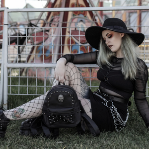 The GothX black vegan skull head mini studded backpack being held by an alternative gothic witchy model with silver hair wearing a gothic styled outfit in front of a fairground ride. The vegan leather bag is facing forward showing off the handle, the skull, mini studs and front zip pocket.