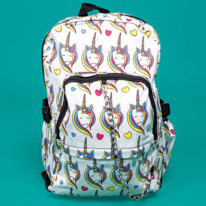 The Rainbow Unicorn Backpack sat facing forward on a teal background. The vegan friendly backpack is a white canvas material with repeating kawaii cute unicorn heads with pink blue and yellow manes with hearts surrounding them. The bag is facing forward to highlight the front two zip pockets, the main zip compartment and silver draping decorative chain.