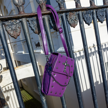 Load image into Gallery viewer, The GothX Purple Mini Coffin Bag hanging off a black metal railing. The coffin bag in a sleek faux leather is a coffin shape with a magnetic close clip flap with a cross chain emblem in the centre, surrounding the coffin shape is mini metal cross studs. Draping across the front of a silver detachable decorative chain.
