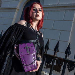 The GothX Purple Mini Coffin Bag being held up by a gothic model wearing a goth black dress with red hair. The coffin bag in a sleek faux leather is a coffin shape with a magnetic close clip flap with a cross chain emblem in the centre, surrounding the coffin shape is mini metal cross studs. Draping across the front of a silver detachable decorative chain with the GothX black cross tag hanging to the left side.