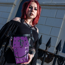 Load image into Gallery viewer, The GothX Purple Mini Coffin Bag being held up by a gothic model wearing a goth black dress with red hair. The coffin bag in a sleek faux leather is a coffin shape with a magnetic close clip flap with a cross chain emblem in the centre, surrounding the coffin shape is mini metal cross studs. Draping across the front of a silver detachable decorative chain with the GothX black cross tag hanging to the left side.
