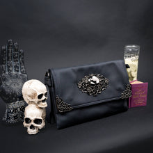 Load image into Gallery viewer, A black vegan leather bag by CHOK GothX with a metallic skull and rose detailing on the front flap, styled with gothic decor including skull props and a spellbook.
