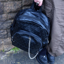 Load image into Gallery viewer, The Black Embossed Cross Backpack being held by a goth grunge model in front of a brick wall in a tumblr grunge style. The backpack is facing forward to highlight the two front silver zip pockets, two elasticated side pockets, main double zip compartment and a silver decorative detachable silver chain draping across the front. The bag is made of vegan friendly leather with 3d embossed crosses in varying sizes all over.
