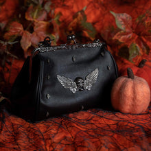 Load image into Gallery viewer, Gothx black vegan winged skull vintage ball clasp handbag on an orange pumpkin background. Bag is facing forward to highlight the ball clasp close, metal detailing, detachable vegan leather handle, mini skull studs and winged skull centre piece.
