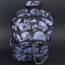 Load image into Gallery viewer, Front view of grey skull print backpack with chain – A gothic backpack featuring a haunting grey skull design on a black background. The front pocket includes a silver chain detail, with one skull showing a striking red eye for a unique, edgy look.
