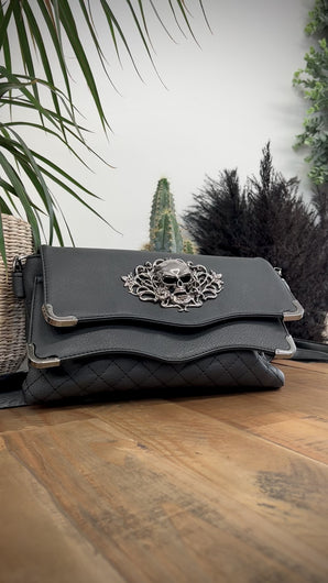 The GothX Skulls and Roses Quilted Clutch Bag sat on a wooden table with black and green plant foliage behind the bag. The vegan leather clutch bag is facing forward and being opened to highlight the two magnetic clip close flaps with metal corners, a stitch quilted front, a skulls and roses metal centrepiece and two D rings on either side for a detachable strap. The mini bag is inspired by gothic grunge witchy fashion.
