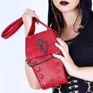 A gothic styled model with dark vampy red makeup wearing a goth black hooded dress holding the GothX Red Mini Coffin Bag to their chest on a white background. The coffin bag is made with vegan friendly sleek red leather and features a detachable decorative metal chain with metal cross studding.