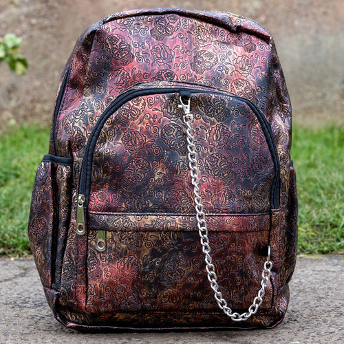 The Rustic Skulls & Roses Backpack sat outside in front of a warm toned grunge wall and grass. The bag is facing forward to highlight the two zip front pockets, two elasticated side pockets, the double zip main compartment with a silver draping chain across the front. The vegan friendly faux leather bag is varying colours of bronze, gold, rose and brown with black 3d embossed sugar skulls, roses and swirls all over.