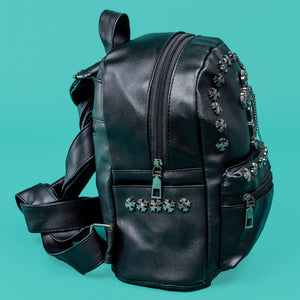 The GothX black cross vegan stud mini backpack on a teal background. The vegan leather bag is facing right to highlight the top handle, adjustable shoulder straps, cross studs along the top zip line and top of zipped pocket, studded cross with chain centrepiece and side metal cross studs on the slot pockets.