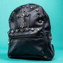 Load image into Gallery viewer, The GothX black cross vegan stud mini backpack on a teal background. The vegan leather bag is facing forwards to highlight the cross studs along the top zip line and top of zipped pocket, studded cross with chain centrepiece and side metal cross studs on the slot pockets.
