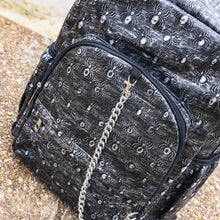 Load image into Gallery viewer, Close up of the Rustic Silver Spider Backpack sat outside on a gritty concrete floor with misty water behind. The bag is facing forward to highlight the two zip front pockets, two elasticated side pockets, the double zip main compartment with a silver draping chain across the front. The vegan friendly faux leather bag has 3d embossed spiders in varying sizes with brushed black and silver tones all over.
