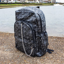 Load image into Gallery viewer, The Rustic Silver Spider Backpack sat outside on a gritty concrete floor with misty water behind. The bag is facing forward angled left to highlight the two zip front pockets, two elasticated side pockets, the double zip main compartment with a silver draping chain across the front. The vegan friendly faux leather bag has 3d embossed spiders in varying sizes with brushed black and silver tones all over.
