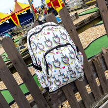 Load image into Gallery viewer, The Rainbow Unicorn Backpack hanging off a wooden fence in front of a multicoloured fairground area by the beach. The vegan friendly backpack is a white canvas material with repeating kawaii cute unicorn heads with pink blue and yellow manes with hearts surrounding them. The bag is facing forward to highlight the front two zip pockets, the main zip compartment and silver draping decorative chain.
