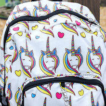 Load image into Gallery viewer, Close up of the Rainbow Unicorn Backpack hanging off a wooden fence in front of a multicoloured fairground area by the beach. The vegan friendly backpack is a white canvas material with repeating kawaii cute unicorn heads with pink blue and yellow manes with hearts surrounding them. The bag is facing forward to highlight the front two zip pockets, the main zip compartment and silver draping decorative chain.
