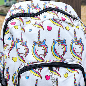 Close up of the Rainbow Unicorn Backpack hanging off a wooden fence in front of a multicoloured fairground area by the beach. The vegan friendly backpack is a white canvas material with repeating kawaii cute unicorn heads with pink blue and yellow manes with hearts surrounding them. The bag is facing forward to highlight the front two zip pockets, the main zip compartment and silver draping decorative chain.