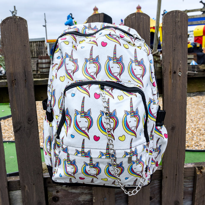 The Rainbow Unicorn Backpack hanging off a wooden fence in front of a multicoloured fairground area by the beach. The vegan friendly backpack is a white canvas material with repeating kawaii cute unicorn heads with pink blue and yellow manes with hearts surrounding them. The bag is facing forward to highlight the front two zip pockets, the main zip compartment and silver draping decorative chain.