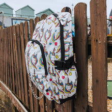 Load image into Gallery viewer, A side view of the Rainbow Unicorn Backpack hanging off a wooden fence in front of a multicoloured fairground area by the beach. The vegan friendly backpack is a white canvas material with repeating kawaii cute unicorn heads with pink blue and yellow manes with hearts surrounding them. The bag is facing forward to highlight the front two zip pockets, the main zip compartment and silver draping decorative chain.
