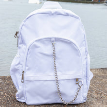 Load image into Gallery viewer, The white backpack with chain is sat outside on concrete floor at a waters edge. The vegan friendly bag is facing forward to highlight the top handle, the double zip main compartment, two front zip pockets with a silver decorative detachable draping chain and two elasticated side pockets.
