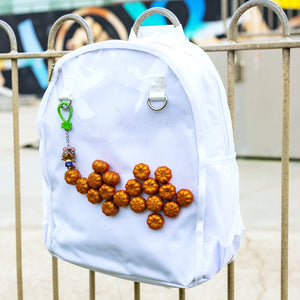 The White & Clear Window Ita Backpack hanging on a metal railing outside a skatepark. The bag is facing forward to highlight the clear front window filled with glittery pumpkins and two metal D rings with a keyring attached, two side elasticated pockets, main zip compartment and top handle. The vegan friendly bag is inspired by kawaii jfashion.