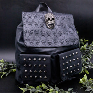 GothX twin pocket skull vegan backpack on a black background with leaves surrounding it. The bag is facing forward to highlight the diamante effect skull, skull embossed vegan leather front flap, tassel tie cords and two silver studded front pockets.