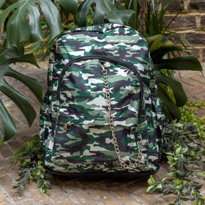 Classic Camouflage vegan backpack with chain sat outside in front of a tropical plant and brick wall. The backpack with a green and brown camo print is facing forward highlighting the two front zip pockets, two side pockets and silver decorative chain.