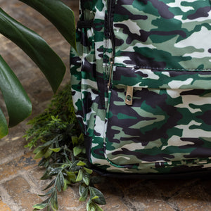 Close up of the Classic Camouflage vegan backpack with chain sat outside in front of a tropical plant and brick wall. The backpack with a green and brown camo print is facing forward highlighting the two front zip pockets, two side pockets and silver decorative chain.