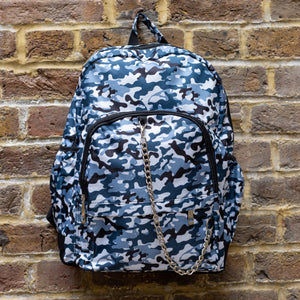 The Snow Camouflage Backpack hanging outside on a brick wall. The white, blue, navy and black vegan friendly backpack is facing forward to highlight the two front zip pockets with a silver draping detachable chain, the two side elasticated pockets, the top handle and the main double zip compartment.