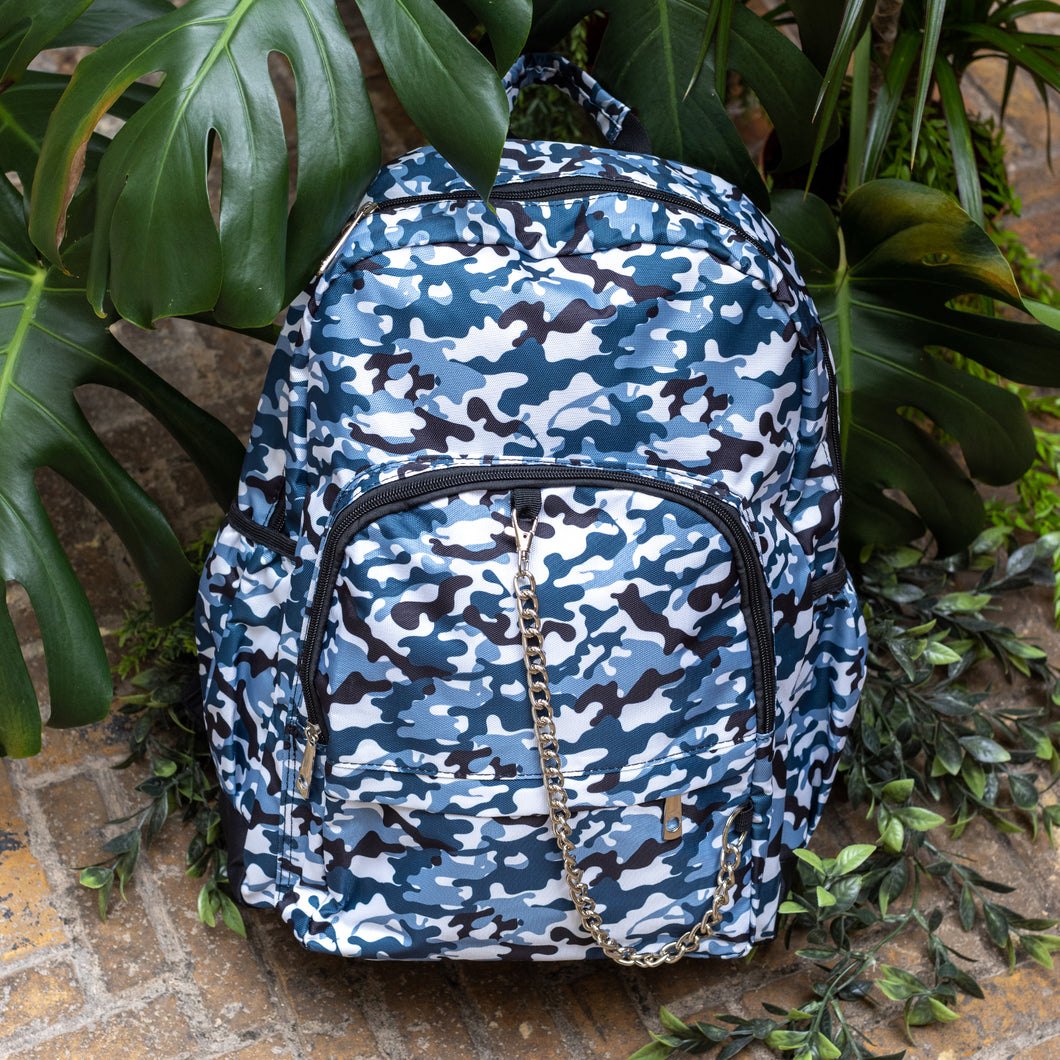 The Snow Camouflage Backpack sat outside on a brick floor surrounded by giant monstera leaves and other green foliage. The white, blue, navy and black vegan friendly camo backpack is sat facing forward to highlight the two front zip pockets with a silver draping detachable chain, the two side elasticated pockets, the top handle and the main double zip compartment.