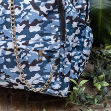 Load image into Gallery viewer, Close up of The Snow Camouflage Backpack sat outside on a brick floor surrounded by giant monstera leaves and other green foliage. The white, blue, navy and black vegan friendly camo backpack is sat facing forward to highlight the two front zip pockets with a silver draping detachable chain, the two side elasticated pockets, the top handle and the main double zip compartment.
