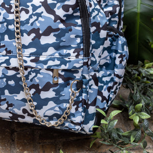 Close up of The Snow Camouflage Backpack sat outside on a brick floor surrounded by giant monstera leaves and other green foliage. The white, blue, navy and black vegan friendly camo backpack is sat facing forward to highlight the two front zip pockets with a silver draping detachable chain, the two side elasticated pockets, the top handle and the main double zip compartment.