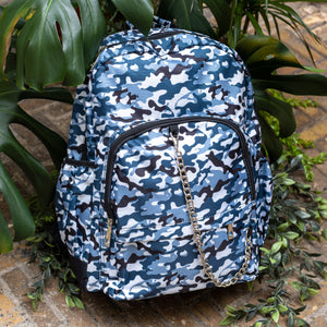 The Snow Camouflage Backpack sat outside on a brick floor surrounded by giant monstera leaves and other green foliage. The white, blue, navy and black vegan friendly camo backpack is sat facing forward to highlight the two front zip pockets with a silver draping detachable chain, the two side elasticated pockets, the top handle and the main double zip compartment.