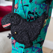 Load image into Gallery viewer, The kawaii black glitter dino vegan bag being worn by a model wearing dino print dungarees. The bag is facing forward to highlight the dinosaur face, moveable arm, detachable strap and black glitter front.
