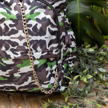 Load image into Gallery viewer, Close up of The Savannah Camouflage Backpack sat outside on a brick floor surrounded by giant monstera leaves and other green foliage. The green, brown, cream, khaki and black vegan friendly backpack is sat facing forward to highlight the two front zip pockets with a silver draping detachable chain, the two side elasticated pockets, the top handle and the main double zip compartment.
