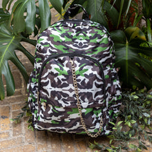 Load image into Gallery viewer, The Savannah Camouflage Backpack sat outside on a brick floor surrounded by giant monstera leaves and other green foliage. The green, brown, cream, khaki and black vegan friendly backpack is sat facing forward to highlight the two front zip pockets with a silver draping detachable chain, the two side elasticated pockets, the top handle and the main double zip compartment.
