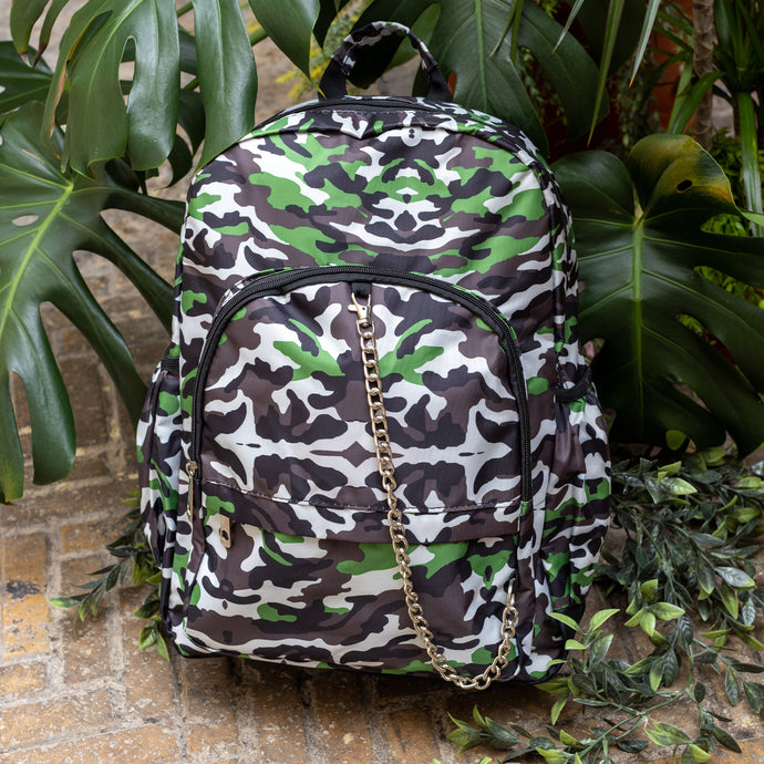 The Savannah Camouflage Backpack sat outside on a brick floor surrounded by giant monstera leaves and other green foliage. The green, brown, cream, khaki and black vegan friendly backpack is sat facing forward to highlight the two front zip pockets with a silver draping detachable chain, the two side elasticated pockets, the top handle and the main double zip compartment.